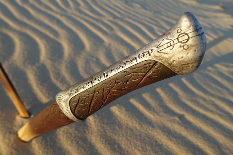 May include: A close-up of a silver and brown metal staff with intricate carvings and a symbol at the top. The staff is lying on a sandy surface.