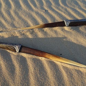 May include: Two decorative knives with wooden handles and silver accents lie on a sandy surface.