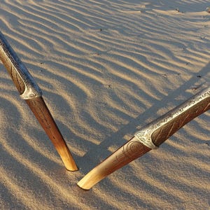 May include: Two ornate silver and brown daggers with intricate designs lie on a sandy surface.