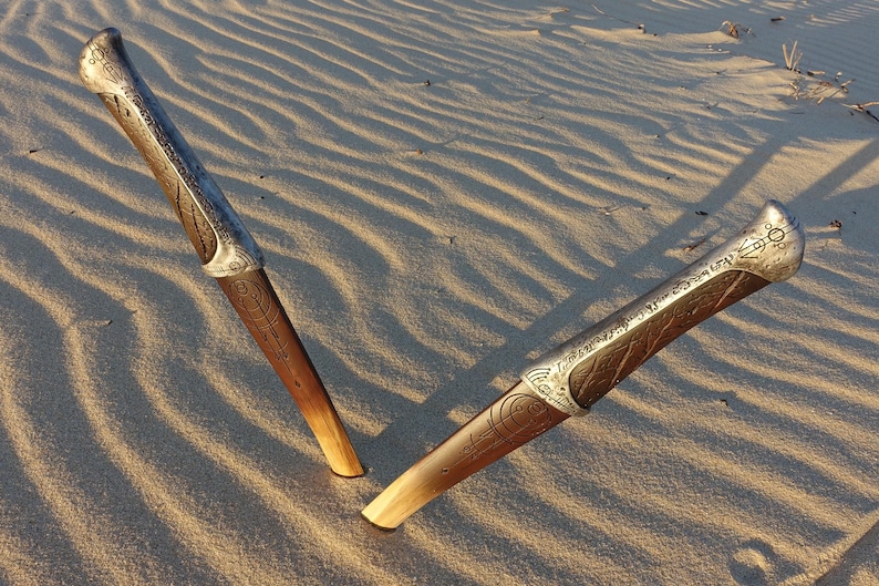 May include: Two ornate silver and brown daggers with intricate designs lie on a sandy surface.