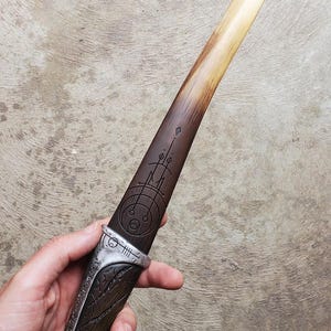 May include: A wooden dagger with a silver handle and a geometric design etched into the blade. The blade is a light brown color and the handle is a darker brown color.