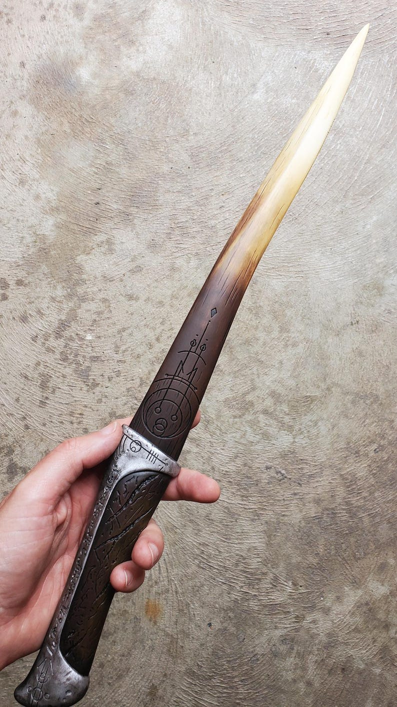 May include: A wooden dagger with a silver handle and a geometric design etched into the blade. The blade is a light brown color and the handle is a darker brown color.