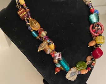 May include: Multi-strand necklace with colorful glass beads, including red, orange, yellow, green, blue, and purple. The necklace features a variety of shapes and sizes of beads, including round, square, and rectangular.