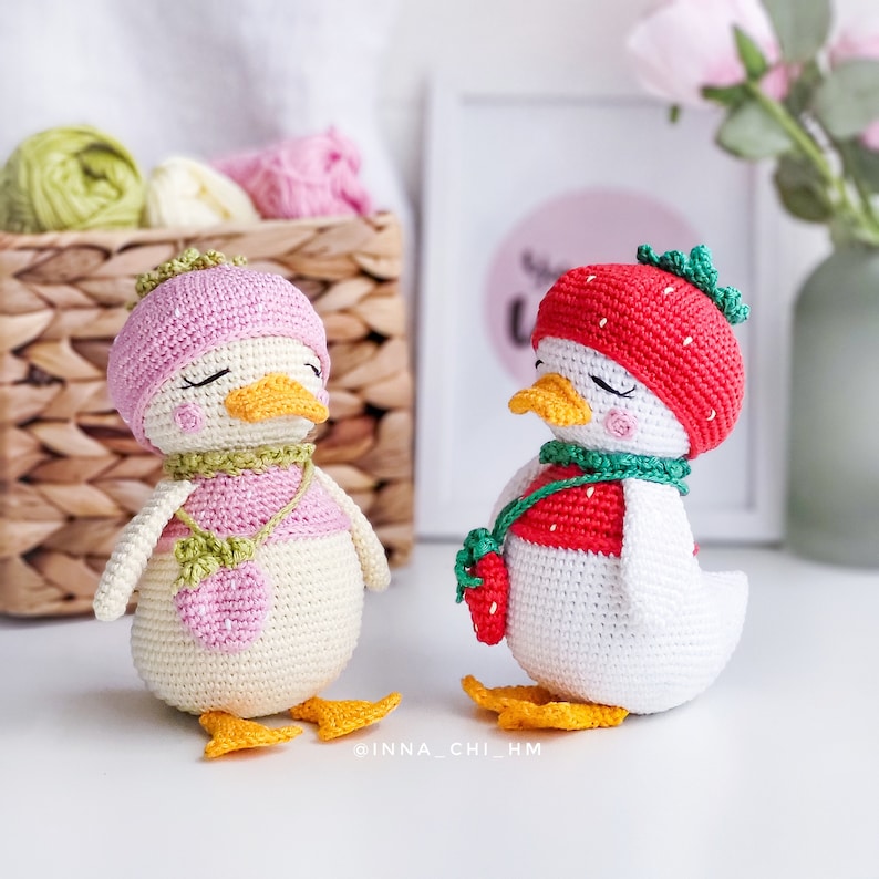 Peut inclure: Deux figurines de canard crochet&eacute;es &agrave; la main. L'un porte un chapeau rose et une &eacute;charpe verte, l'autre un chapeau fraise rouge et une &eacute;charpe verte. Les deux ont des becs et des pieds jaunes et les yeux ferm&eacute;s. Les canards sont sur une surface blanche.