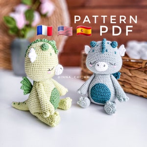 以下が含まれることがあります： 緑とグレーの2つの編みぐるみドラゴンのおもちゃ。グレーのドラゴンは青いアクセントが付いています。おもちゃは白い表面の上に置かれています。画像には「PATTERN PDF」というテキストと、フランス、アメリカ、スペインの国旗が含まれています。