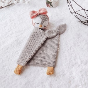 Peut inclure: Une couverture b&eacute;b&eacute; en crochet gris avec un jouet en crochet de pingouin sur le dessus. Le pingouin a un n&oelig;ud rose et un visage blanc avec des yeux noirs et un bec jaune. La couverture a une queue en crochet et deux pieds en crochet.