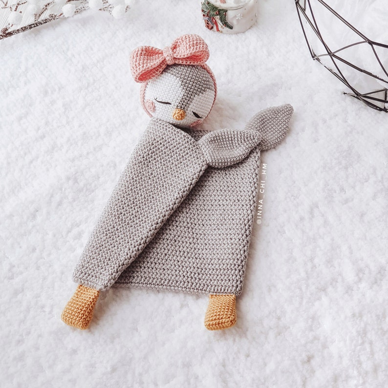 Peut inclure: Une couverture b&eacute;b&eacute; en crochet gris avec un jouet en crochet de pingouin sur le dessus. Le pingouin a un n&oelig;ud rose et un visage blanc avec des yeux noirs et un bec jaune. La couverture a une queue en crochet et deux pieds en crochet.