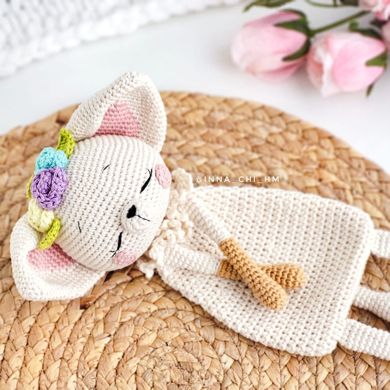 May include: A crocheted white bunny with pink ears and a flower crown is lying on a woven straw mat. The bunny has a brown crocheted nose and closed eyes.