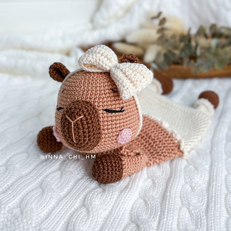 May include: A crocheted stuffed animal resembling a llama with a white bow, lying on a white knitted blanket. The llama has brown eyes, a brown nose, and pink cheeks.