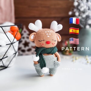 Puede incluir: Un juguete de reno de crochet con una bufanda verde y un mono gris. El reno tiene una nariz roja y cuernos blancos. El juguete está sentado sobre una superficie blanca. La imagen también incluye el texto "PATTERN" y "@INNA_CHI_HM".