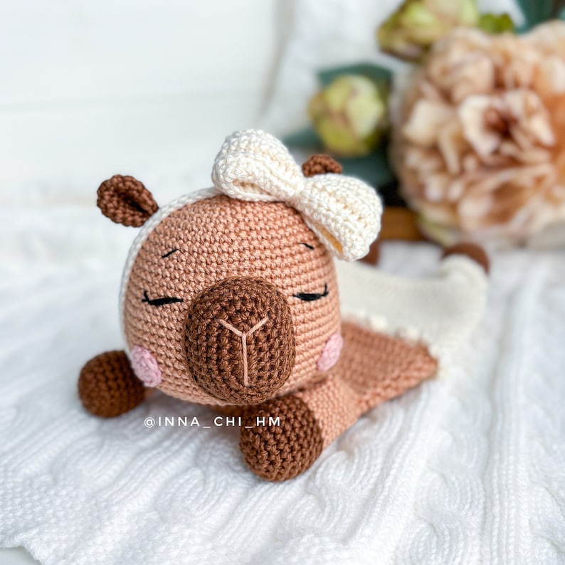 May include: A crocheted brown and white llama toy with a white bow on its head. The llama is lying down with its eyes closed. The toy is on a white knitted blanket.