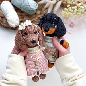 May include: Two crocheted dachshund dogs. One is brown with a pink dress and a flower crown. The other is black and tan, wearing a striped shirt and overalls. The text "INNA CHI HM" is visible on the pink dress.