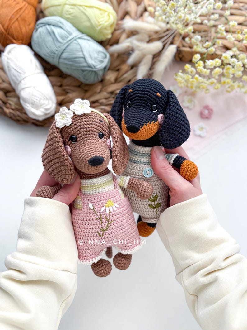 May include: Two crocheted dachshund dogs. One is brown with a pink dress and a flower crown. The other is black and tan, wearing a striped shirt and overalls. The text "INNA CHI HM" is visible on the pink dress.