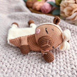 May include: A crocheted brown and white stuffed animal with a pink nose and a white bow. The animal is lying on a pink knitted blanket.