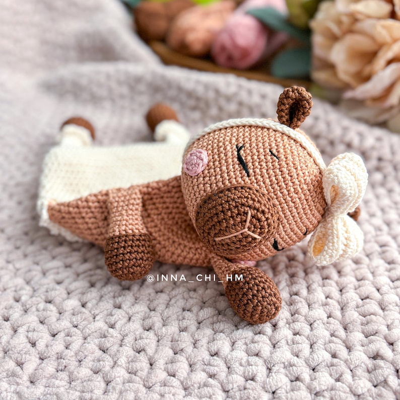 May include: A crocheted brown and white stuffed animal with a pink nose and a white bow. The animal is lying on a pink knitted blanket.