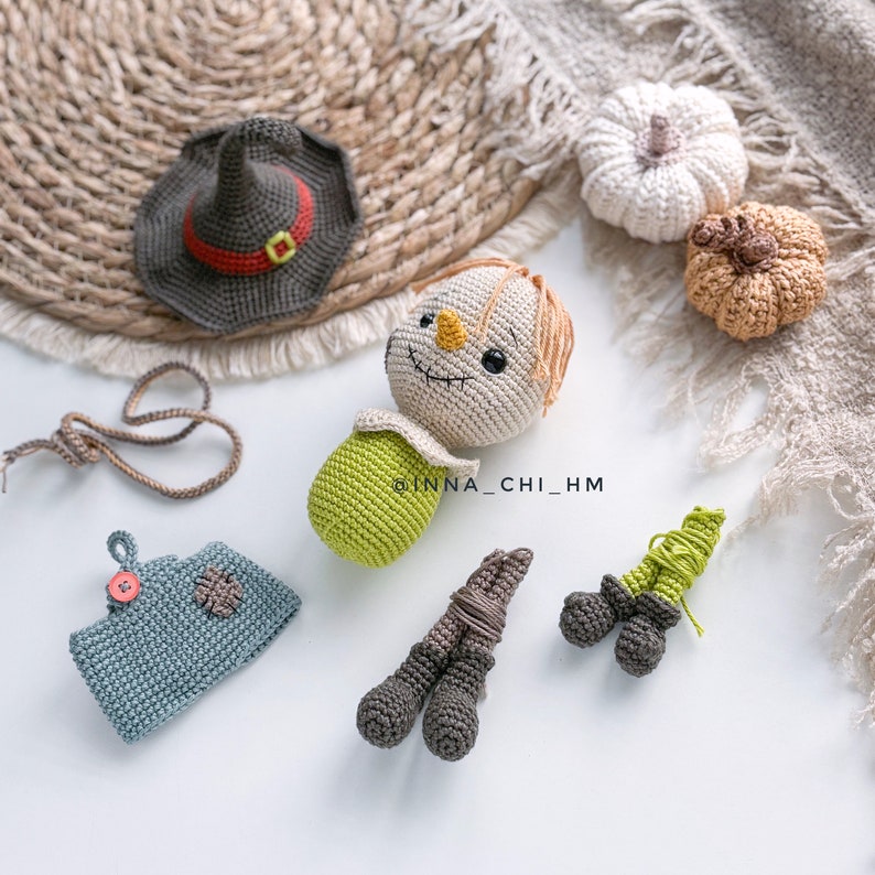 Op de afbeelding: Een verzameling handgemaakte herfstthema-decoraties van gehaakt garen. Inclusief een vogelverschrikkerpop, een heksenhoed, twee pompoenen, een klein blauw zakje en twee sets benen. De vogelverschrikker heeft een beige gezicht, oranje haar en een groene romp.