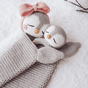 Peut inclure: Deux jouets en forme de pingouin en crochet, l'un avec un n&oelig;ud rose, blottis l'un contre l'autre dans une couverture en crochet grise. La couverture porte le texte "@inna_chi_hm" dans le coin inf&eacute;rieur droit.
