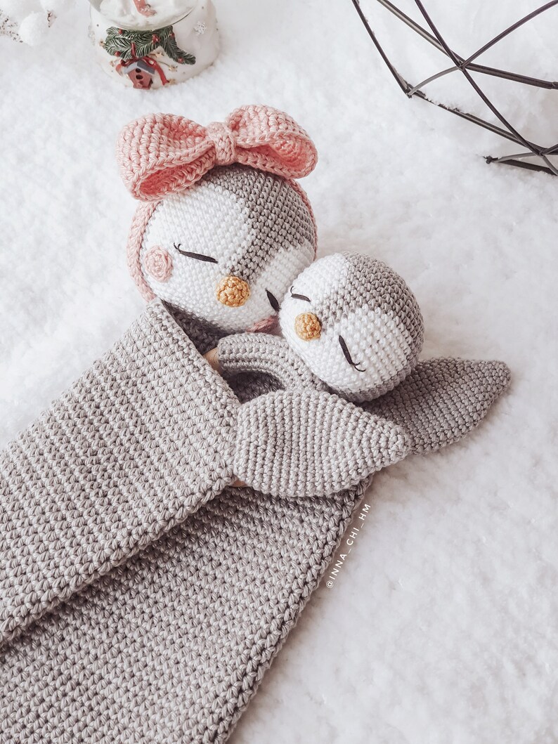 Peut inclure: Deux jouets en forme de pingouin en crochet, l'un avec un n&oelig;ud rose, blottis l'un contre l'autre dans une couverture en crochet grise. La couverture porte le texte "@inna_chi_hm" dans le coin inf&eacute;rieur droit.