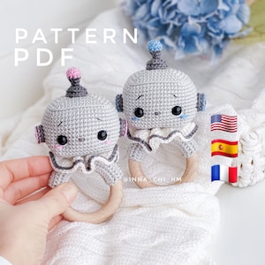 Könnte beinhalten: Zwei gehäkelte Roboter-Rasselspielzeuge mit Holzringen. Ein Roboter ist grau mit einer rosa Oberseite und der andere ist grau mit einer blauen Oberseite. Die Roboter haben schwarze Augen und einen kleinen rosa Mund. Die Spielzeuge liegen auf einer weißen Strickdecke.