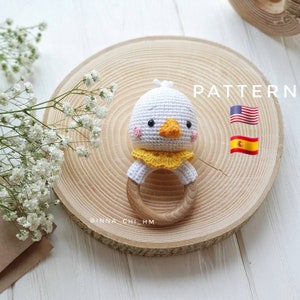 May include: A white crocheted baby rattle with a yellow ruffle and a cute chick face. The rattle is attached to a wooden ring. The image includes the text "PATTERN" and flags for the United States and Spain.
