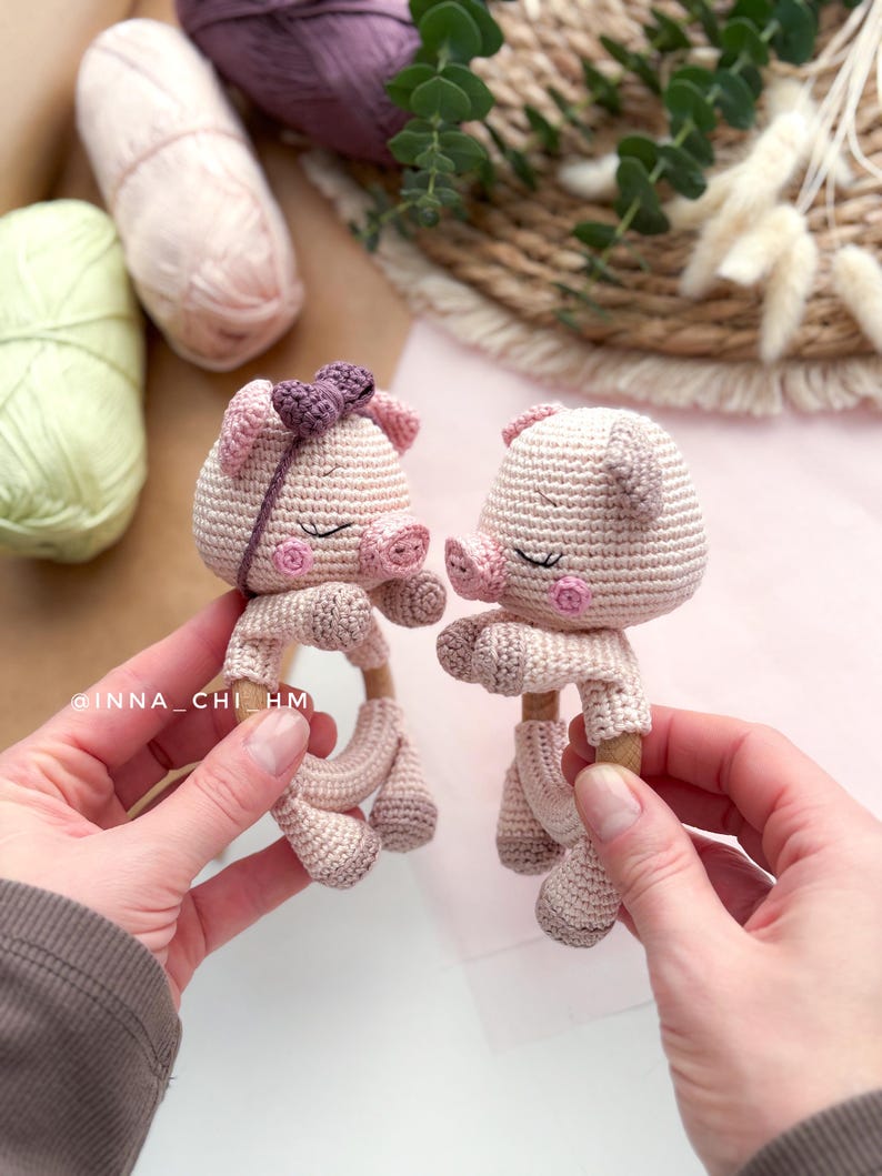 May include: Two handmade crochet piglet baby rattles in light pink, held by hands. Each pig has a round head, small ears, closed eyes, and a wooden ring base. One piglet has a purple bow. The background includes yarn and greenery.