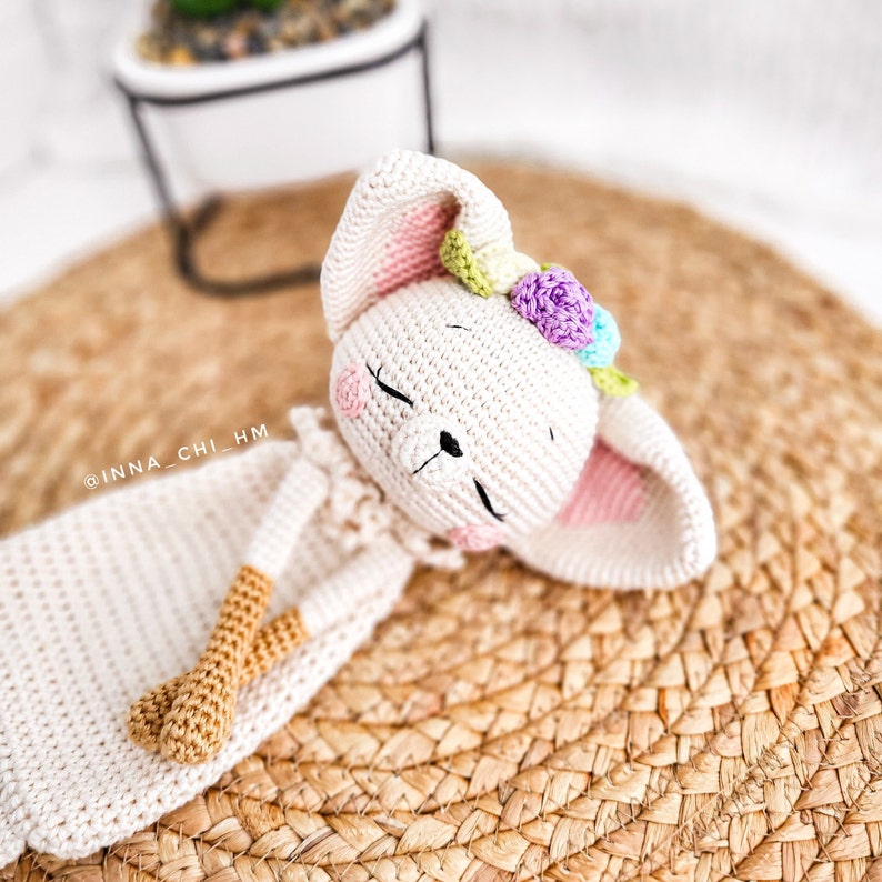 May include: A crocheted white bunny with pink ears and a flower crown. The bunny is lying down with its eyes closed. The bunny is on a woven straw mat.