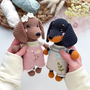 May include: Two handmade crochet dachshund dogs. One is brown with a pink dress and flower crown, the other is black and tan with overalls. The dogs are held in front of a background with yarn and flowers. The text "INNA_CHI_HM" is visible.
