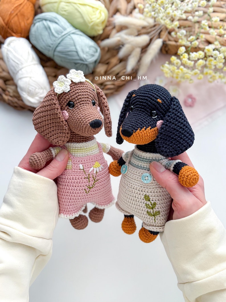 May include: Two handmade crochet dachshund dogs. One is brown with a pink dress and flower crown, the other is black and tan with overalls. The dogs are held in front of a background with yarn and flowers. The text "INNA_CHI_HM" is visible.