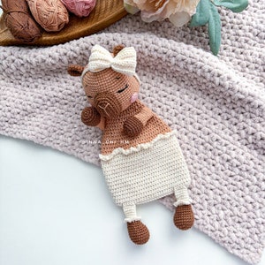 May include: A crocheted brown and white llama toy with a white bow. The llama is wearing a white crocheted dress and has a white crocheted blanket.
