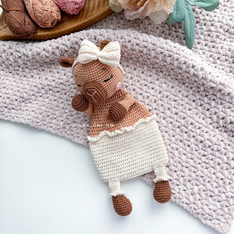 May include: A crocheted brown and white llama toy with a white bow. The llama is wearing a white crocheted dress and has a white crocheted blanket.
