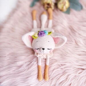 May include: White crocheted stuffed animal with pink ears and a flower crown. The animal has closed eyes and a small black nose. The animal is lying on a pink fluffy surface.