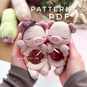 Puede incluir: Dos mordedores de bebé de cerdito de ganchillo. Los mordedores de cerdito son de color rosa claro con detalles en rosa oscuro y un anillo de madera. Un cerdito tiene un lazo morado. La imagen incluye el texto "PATTERN PDF" y la bandera americana.