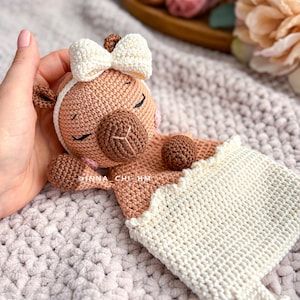 May include: A crocheted brown and white llama toy with a white bow, sleeping in a white crocheted blanket. The toy has a brown nose and closed eyes.