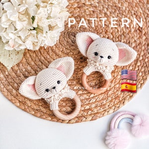 Puede incluir: Dos juguetes de sonajero de cachorro blanco de crochet con orejas rosas y anillos de madera. Los juguetes están sobre una superficie de mimbre marrón con la palabra "PATTERN" en blanco arriba.