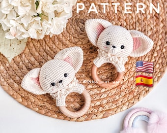 May include: Two crocheted white puppy dog rattle toys with pink ears and wooden rings. The toys are on a woven brown surface with the word "PATTERN" in white text above them.