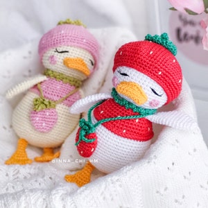 Peut inclure: Deux poup&eacute;es canard crochet&eacute;es &agrave; la main. L'une porte un chapeau rose et une &eacute;charpe verte, l'autre un chapeau et un gilet fraise rouges. Les deux ont les yeux ferm&eacute;s, un bec orange et des pieds jaunes. Les poup&eacute;es reposent sur une surface blanche textur&eacute;e.