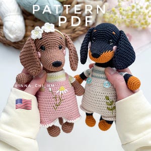Può includere: Due bambole di bassotto all'uncinetto. Una è marrone con un vestito rosa e dettagli a margherita, l'altra è nera e marrone chiaro con un vestito beige. Le bambole sono tenute davanti a un cestino con filato e la scritta "PATTERN PDF".