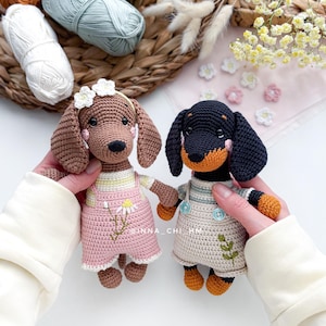 May include: Two crocheted dachshund dog dolls. One is brown with a pink outfit and a daisy detail. The other is black and tan with a beige outfit. Both dolls have button details and are handmade. The background includes yarn and flowers.
