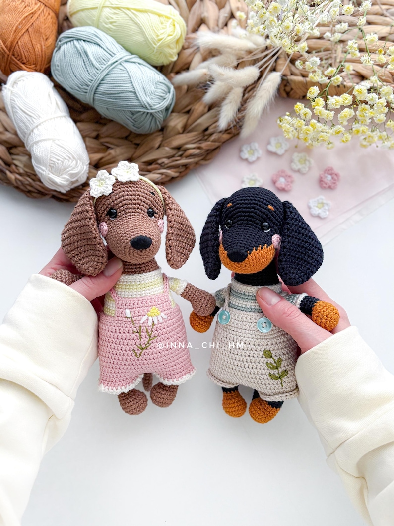 May include: Two crocheted dachshund dog dolls. One is brown with a pink outfit and a daisy detail. The other is black and tan with a beige outfit. Both dolls have button details and are handmade. The background includes yarn and flowers.