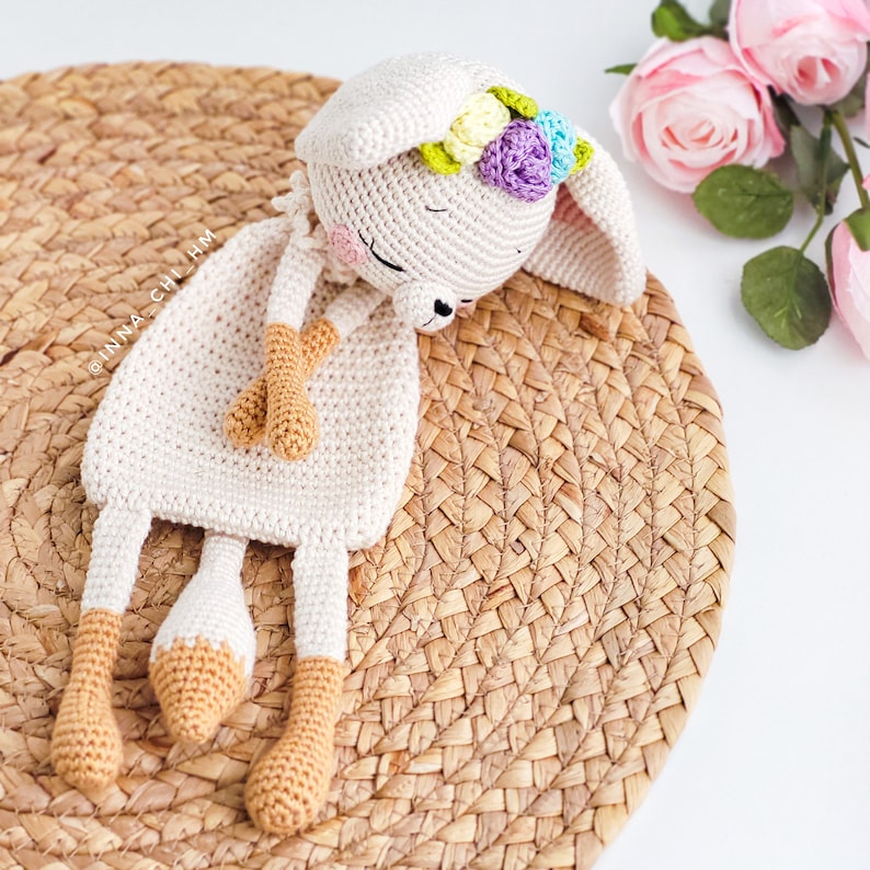 May include: A crocheted bunny toy with a white body and orange legs. The bunny is wearing a flower crown with purple, blue, yellow, and green flowers. The bunny is lying on a woven straw mat.