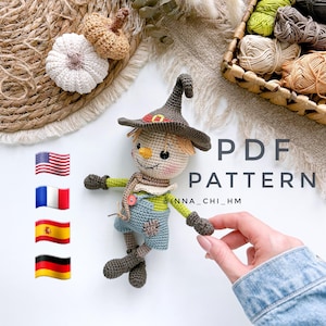 以下が含まれることがあります： 茶色の帽子、麦わら色の髪、青いオーバーオールを着た、手作りのかぎ針編みのカカシ人形。人形には縫い付けられた顔があり、小さなボタンを持っています。画像には「PDF PATTERN」というテキストも含まれています。