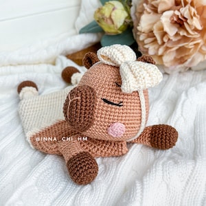 May include: A crocheted brown and white pig with a white bow on its head. The pig is lying on a white knitted blanket. The pig has a pink nose and brown eyes.