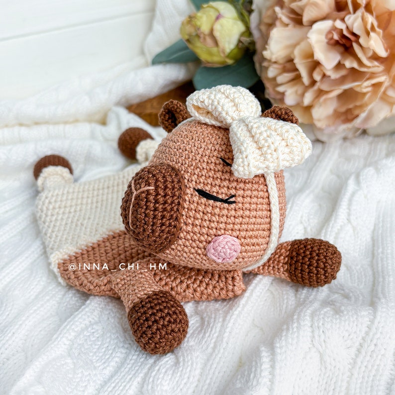 May include: A crocheted brown and white pig with a white bow on its head. The pig is lying on a white knitted blanket. The pig has a pink nose and brown eyes.