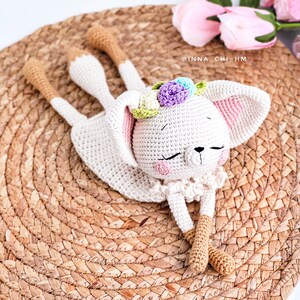 May include: A white crocheted bunny with closed eyes, wearing a flower crown with pink, purple and blue flowers. The bunny is lying on a woven straw mat. The text '@INNA_CHI_HM' is visible in the background.