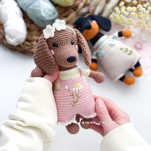 May include: A crocheted dachshund dog doll with brown fur, wearing pink overalls with a daisy design and a white flower crown. Another crocheted dog is in the background. The image includes the text "@INNA_CHI_HM".
