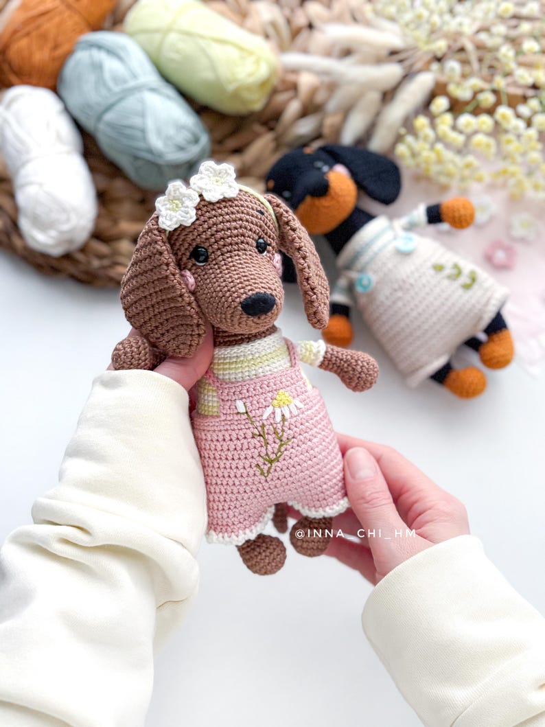 May include: A crocheted dachshund dog doll with brown fur, wearing pink overalls with a daisy design and a white flower crown. Another crocheted dog is in the background. The image includes the text "@INNA_CHI_HM".