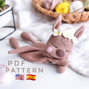 Puede incluir: Un juguete de amigurumi de canguro tejido a crochet con los ojos cerrados, una nariz rosa y una corona de flores blancas. El juguete está acostado sobre una manta blanca con el texto "PDF PATTERN" y las banderas de Estados Unidos y España.