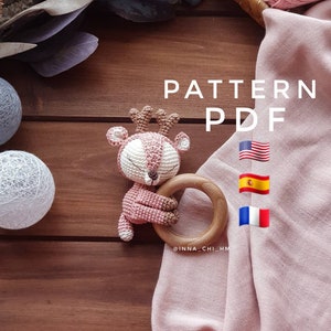 Puede incluir: Un juguete de crochet de ciervo rosa con los ojos cerrados sosteniendo un anillo de dentición de madera. El juguete está sobre una superficie de madera con una manta rosa y el texto "PATTERN PDF" en blanco. Las banderas de Estados Unidos, España y Francia también son visibles.