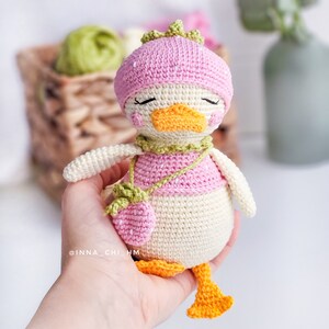 Peut inclure: Une poup&eacute;e canard au crochet faite &agrave; la main, avec un chapeau rose, une chemise et un sac &agrave; main en forme de fraise. Le canard a un bec et des pieds oranges, avec une &eacute;charpe verte. La poup&eacute;e est de couleur cr&egrave;me et a les yeux ferm&eacute;s.