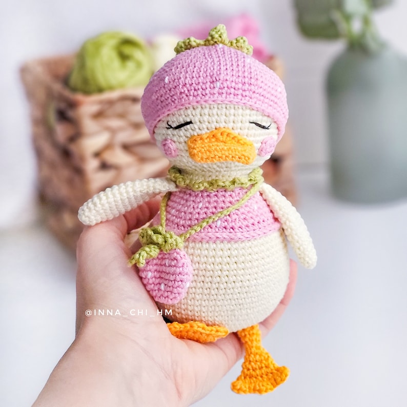 Peut inclure: Une poup&eacute;e canard au crochet faite &agrave; la main, avec un chapeau rose, une chemise et un sac &agrave; main en forme de fraise. Le canard a un bec et des pieds oranges, avec une &eacute;charpe verte. La poup&eacute;e est de couleur cr&egrave;me et a les yeux ferm&eacute;s.